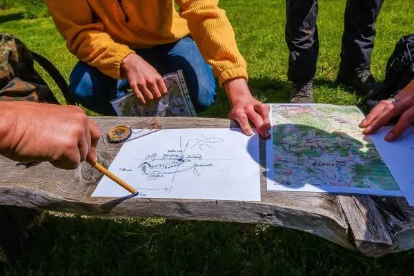 Karte und Kompass liegen auf einer Holzbank, Hände zeigen auf die Zeichnungen